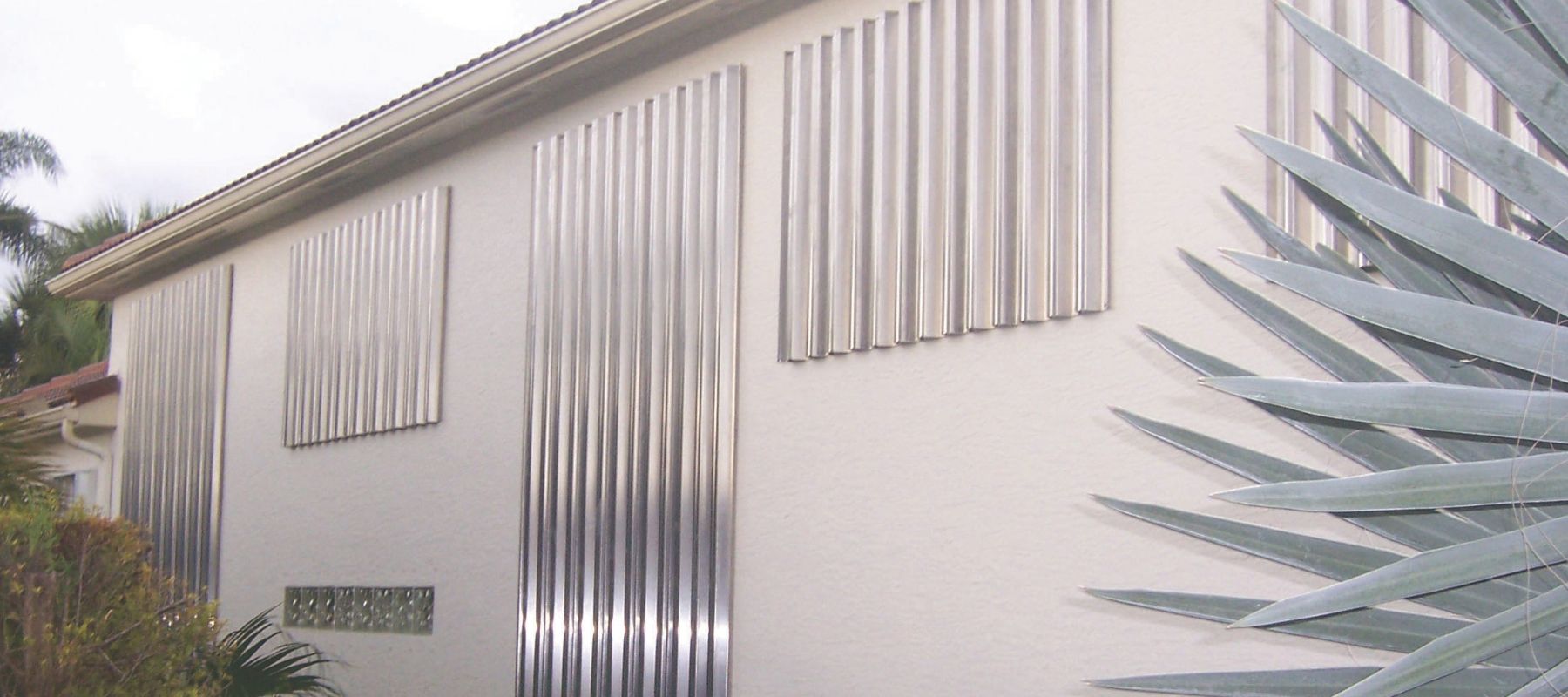 Corrugated metal hurricane shutters cover windows on a white stucco home